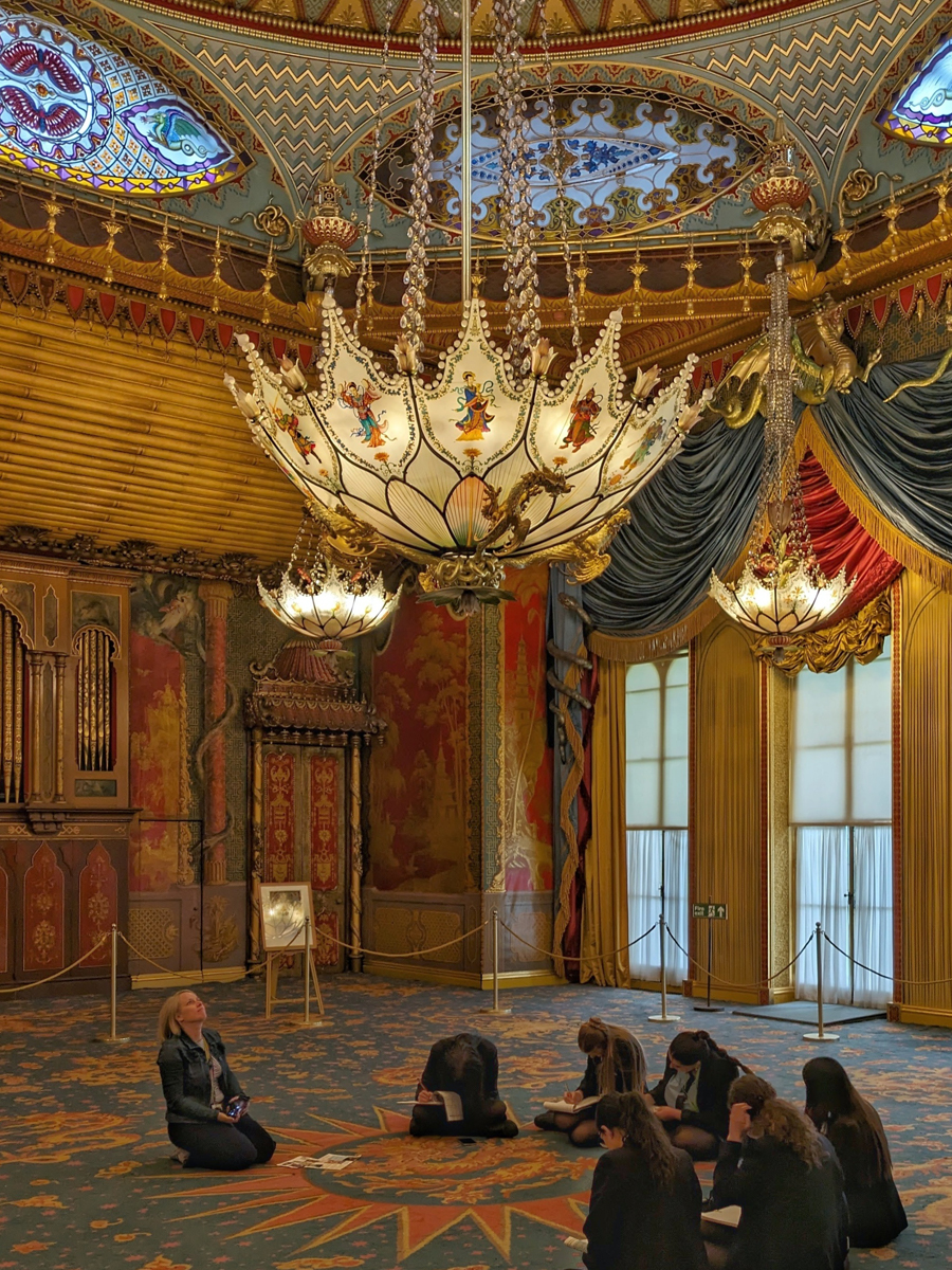 Drawing Dragons, a KS3 & KS4 school group session in the Royal Pavilion. A group of school children sit on the floor in the Music Room of the Royal Pavilion. They have A4 notepads and pencils and are drawing. The teacher sits with them, looking up at the ceiling.