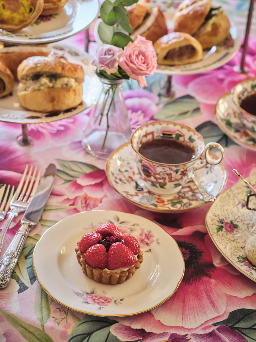 Afternoon tea in the Adelaide Suite of the Royal Pavilion. A close up of the table with a cup of tea on a saucer and a plate with a strawberry tart. A small vase of flowers and a plate of scones are in the background.