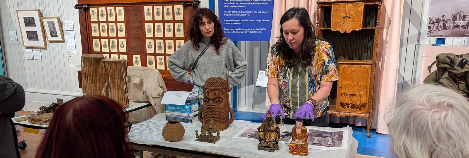 Curator at Work - Portia Tremlett. Portia stands behind a table in the Central Gallery of Brighton Museum. Objects from the World Art collection are on the table and Portia holds a Chinese Deity object as she speaks