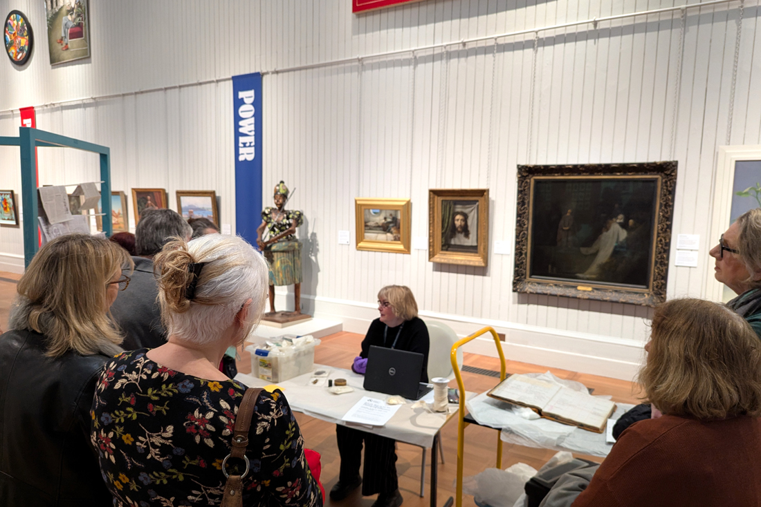 Curator at Work - Lucy Faithful. Lucy sits behind a desk in the Central Gallery of Brighton Museum. On the desk is a laptop and a plastic storage box containing various wrapped objects. A group of people stand facing the desk at Lucy unwraps and talks about the objects.