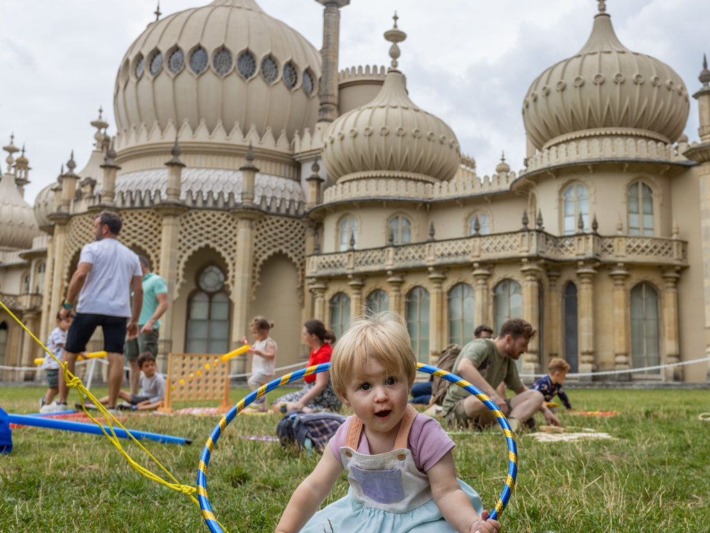 Photo of the family event for Pride, Picnic Like A Prince in the grounds of the Royal Pavilion. Groups of families sit on the lawn playing games. A young girl plays with a hula hoop in the foreground