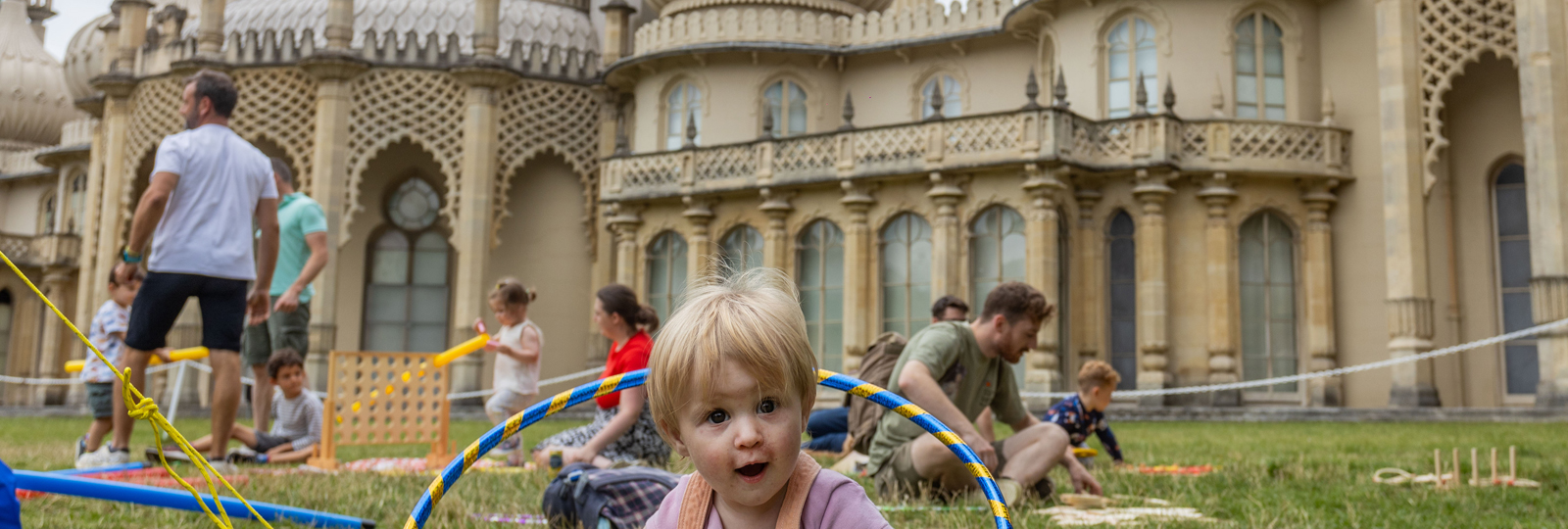 Photo of the family event for Pride, Picnic Like A Prince in the grounds of the Royal Pavilion. Groups of families sit on the lawn playing games. A young girl plays with a hula hoop in the foreground