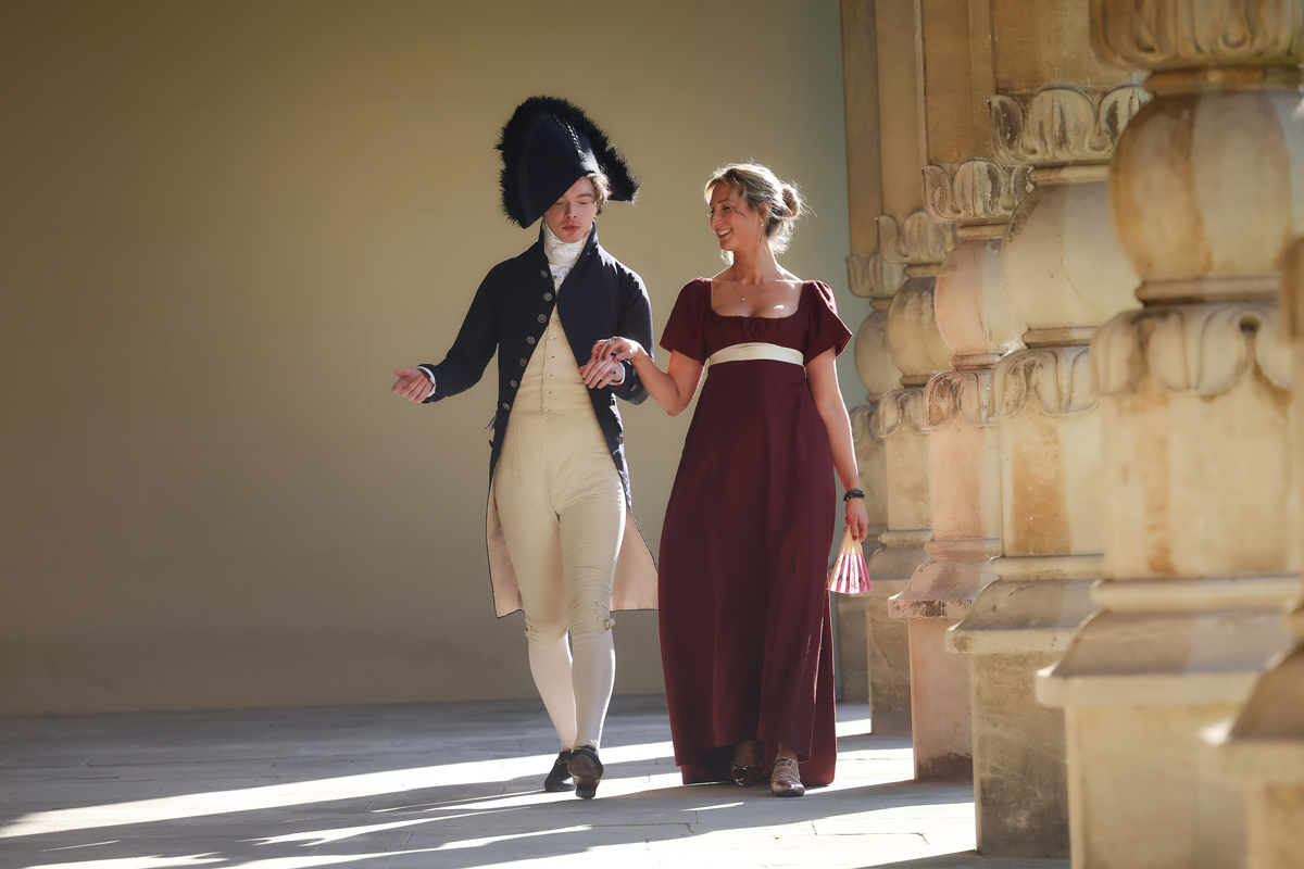 Regency couple photoshoot at the Royal Pavilion. A man and woman in Regency dress are walking along the outside of the Royal Pavilion. She wears a dark red dress with a white ribbon belt. He wears cream trousers, blouse and waistcoat with a navy long coat and large circular navy hat