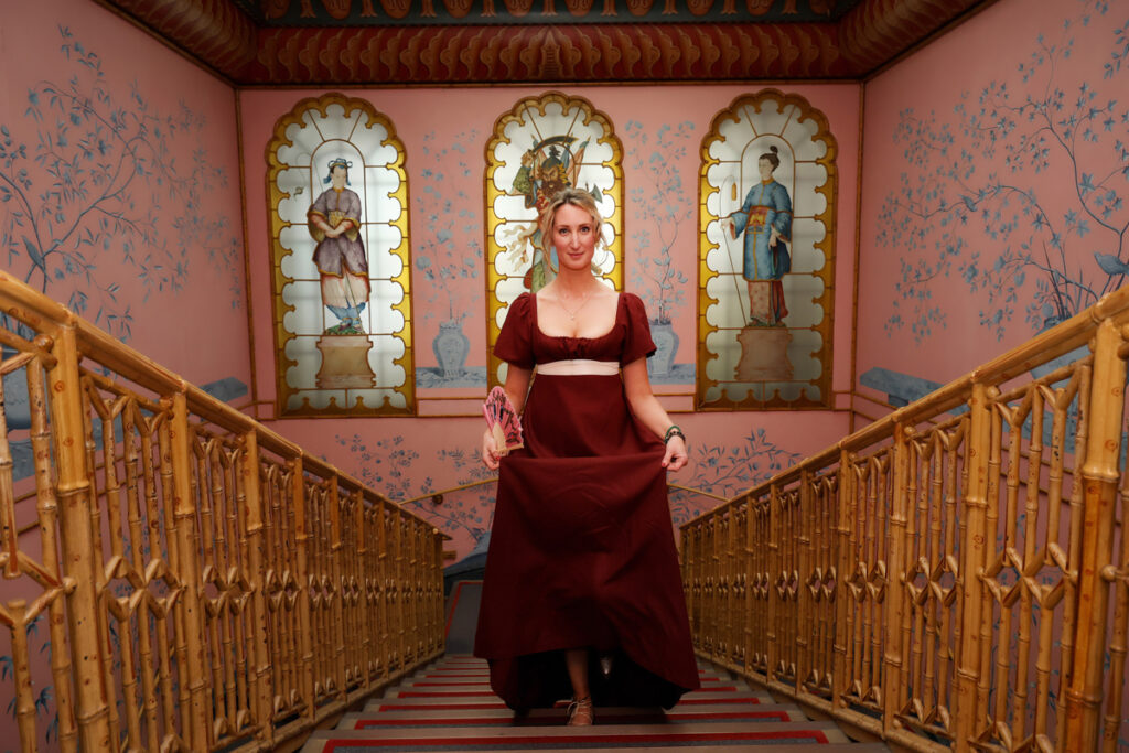 Regency couple photoshoot at the Royal Pavilion. A woman in Regency dress is walking up the stairs in the Royal Pavilion. She wears a dark red dress with a white ribbon belt.