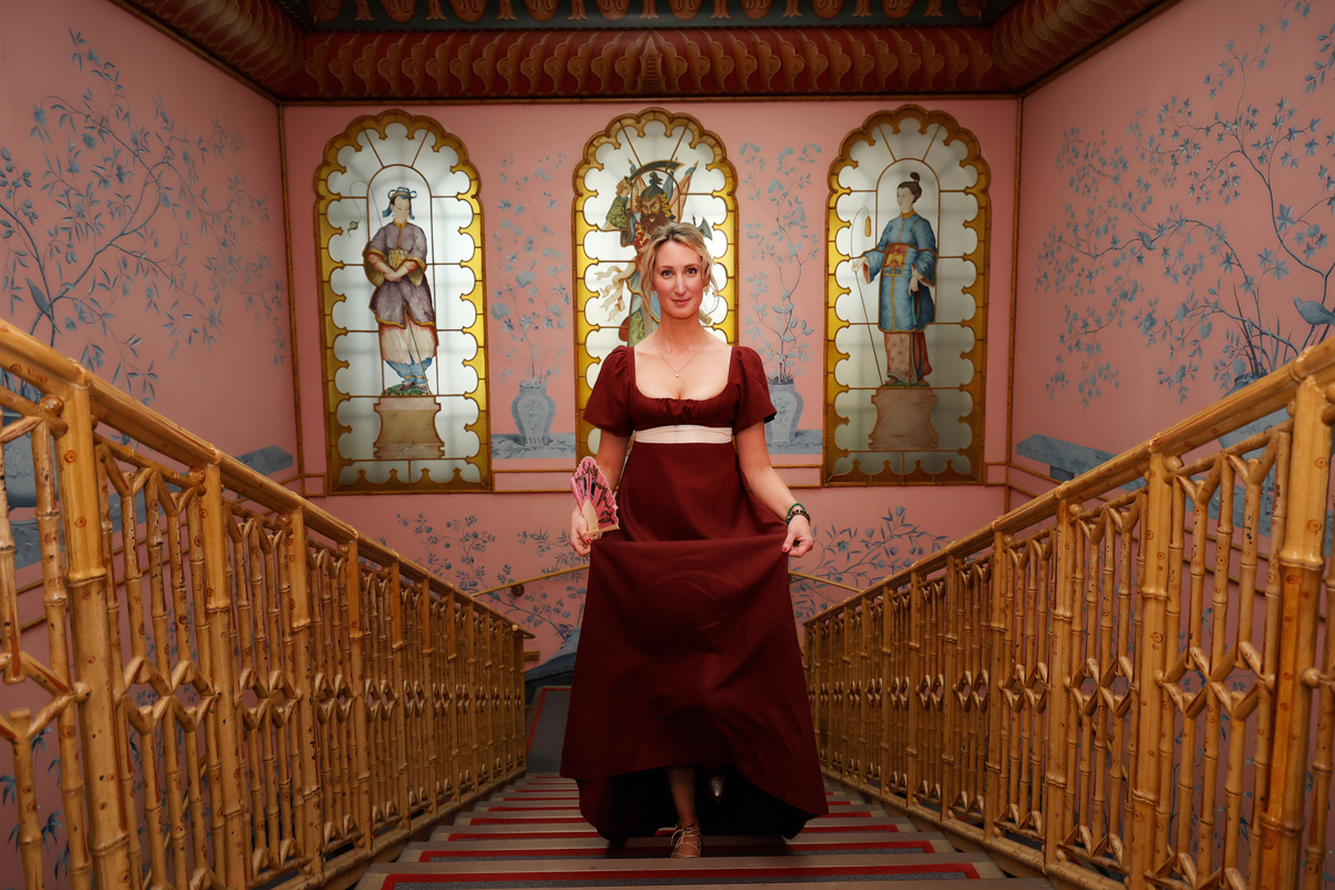 Regency couple photoshoot at the Royal Pavilion. A woman in Regency dress is walking up the stairs in the Royal Pavilion. She wears a dark red dress with a white ribbon belt.