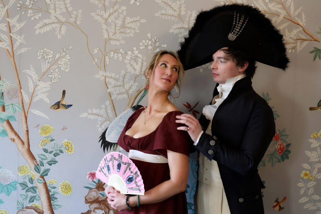 Regency couple photoshoot at the Royal Pavilion. A man and woman in Regency dress are in the William IV Room. She wears a dark red dress with a white ribbon belt. He wears cream trousers, blouse and waistcoat with a navy long coat and large circular navy hat