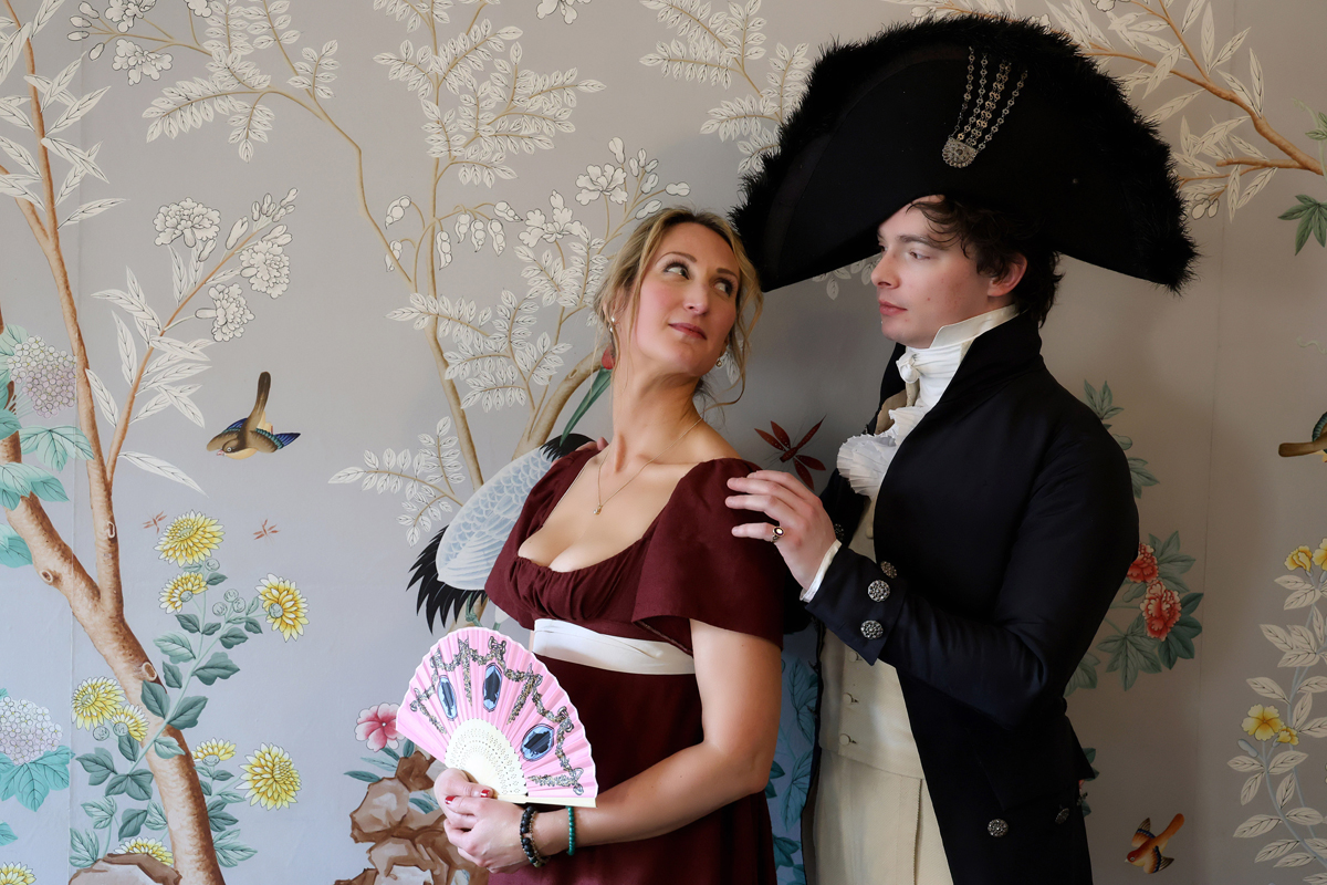 Regency couple photoshoot at the Royal Pavilion. A man and woman in Regency dress are in the William IV Room. She wears a dark red dress with a white ribbon belt. He wears cream trousers, blouse and waistcoat with a navy long coat and large circular navy hat