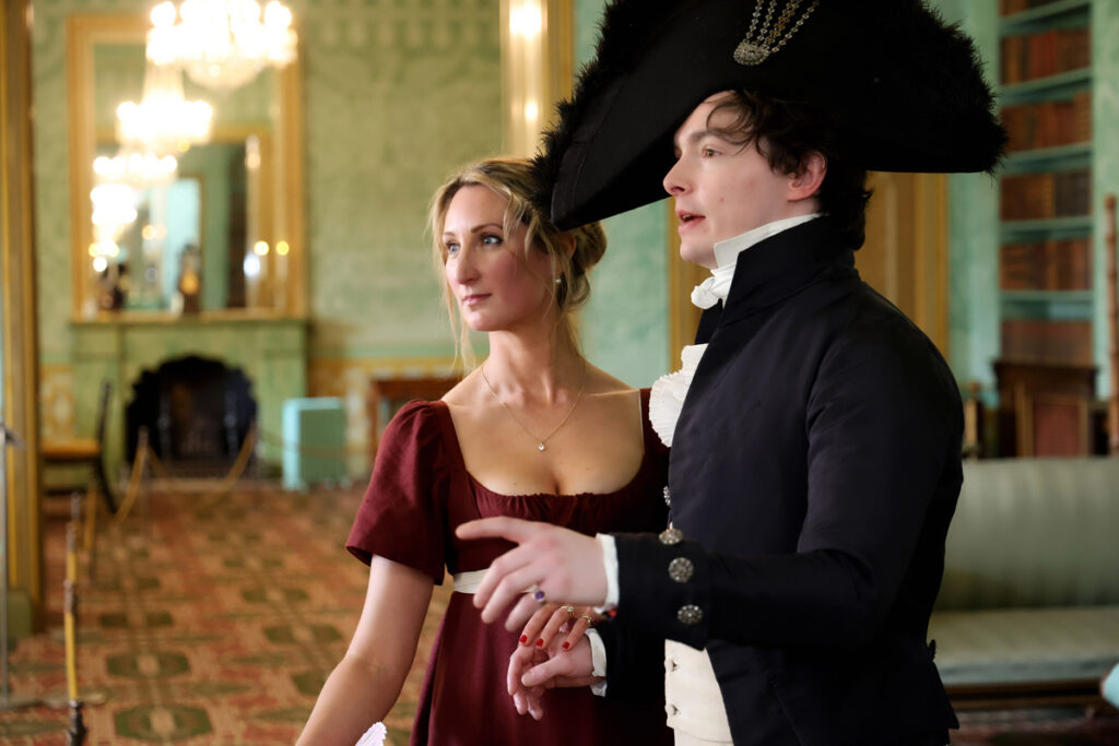 Regency couple photoshoot at the Royal Pavilion. A man and woman in Regency dress are in the King's Apartment. She wears a dark red dress with a white ribbon belt. He wears cream trousers, blouse and waistcoat with a navy long coat and large circular navy hat