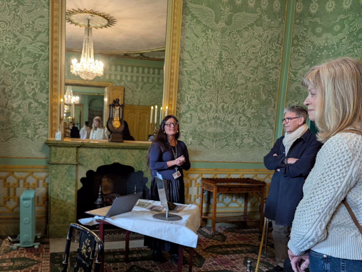 Curator at Work - Saadia. Saadia stands in the King's Apartments of the Royal Pavilion talking to a group of people. She stands by her work table which has a laptop and printed archives