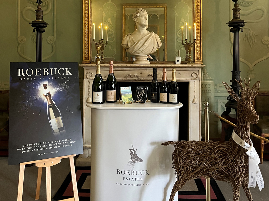 Roebuck Estates wine promotional image inside the Royal Pavilion. A temporary bar plinth with bottles of wine is placed in front of a fireplace. A bust of King George IV sits on the mantle above. To the left of the bar is a Roebuck Wine poster on a wooden easel. A wicker sculpture of a deer - the Roebuck logo - is placed to the right of the bar.