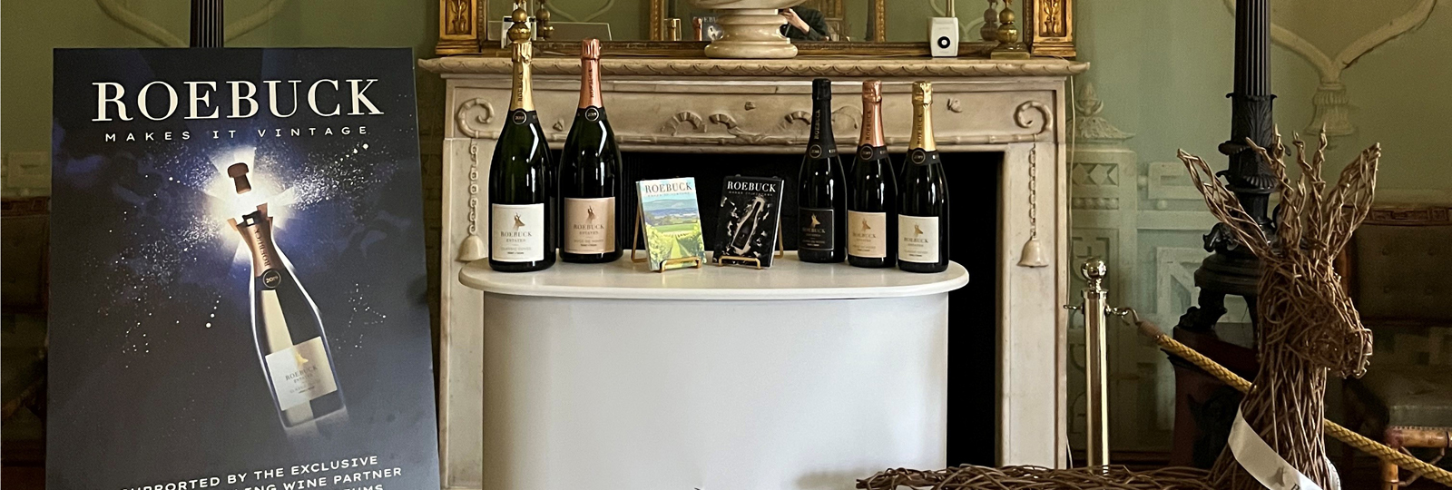 Roebuck Estates wine promotional image inside the Royal Pavilion. A temporary bar plinth with bottles of wine is placed in front of a fireplace. A bust of King George IV sits on the mantle above. To the left of the bar is a Roebuck Wine poster on a wooden easel. A wicker sculpture of a deer - the Roebuck logo - is placed to the right of the bar.