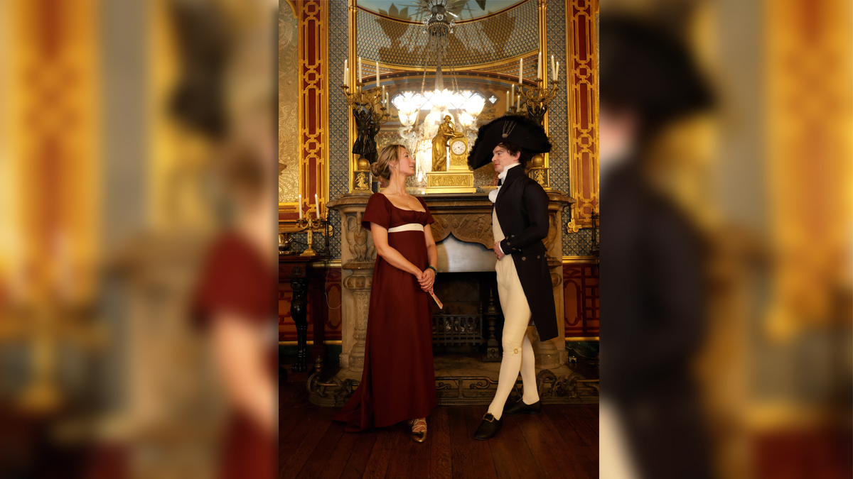 Regency couple photoshoot at the Royal Pavilion. A man and woman in Regency dress stand in front of a fireplace. She wears a dark red dress with a white ribbon belt. He wears cream trousers, blouse and waistcoat with a navy long coat and large circular navy hat