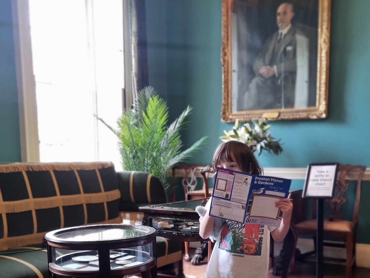 Preston Manor Easter trail 2026. A child stands in the entry hall of Preston Manor. Their face is hidden behind a printed Preston Manor children's trail leaflet they hold in one hand, the other hand holds a magnifying glass and a toy bunny is tucked under their arm.