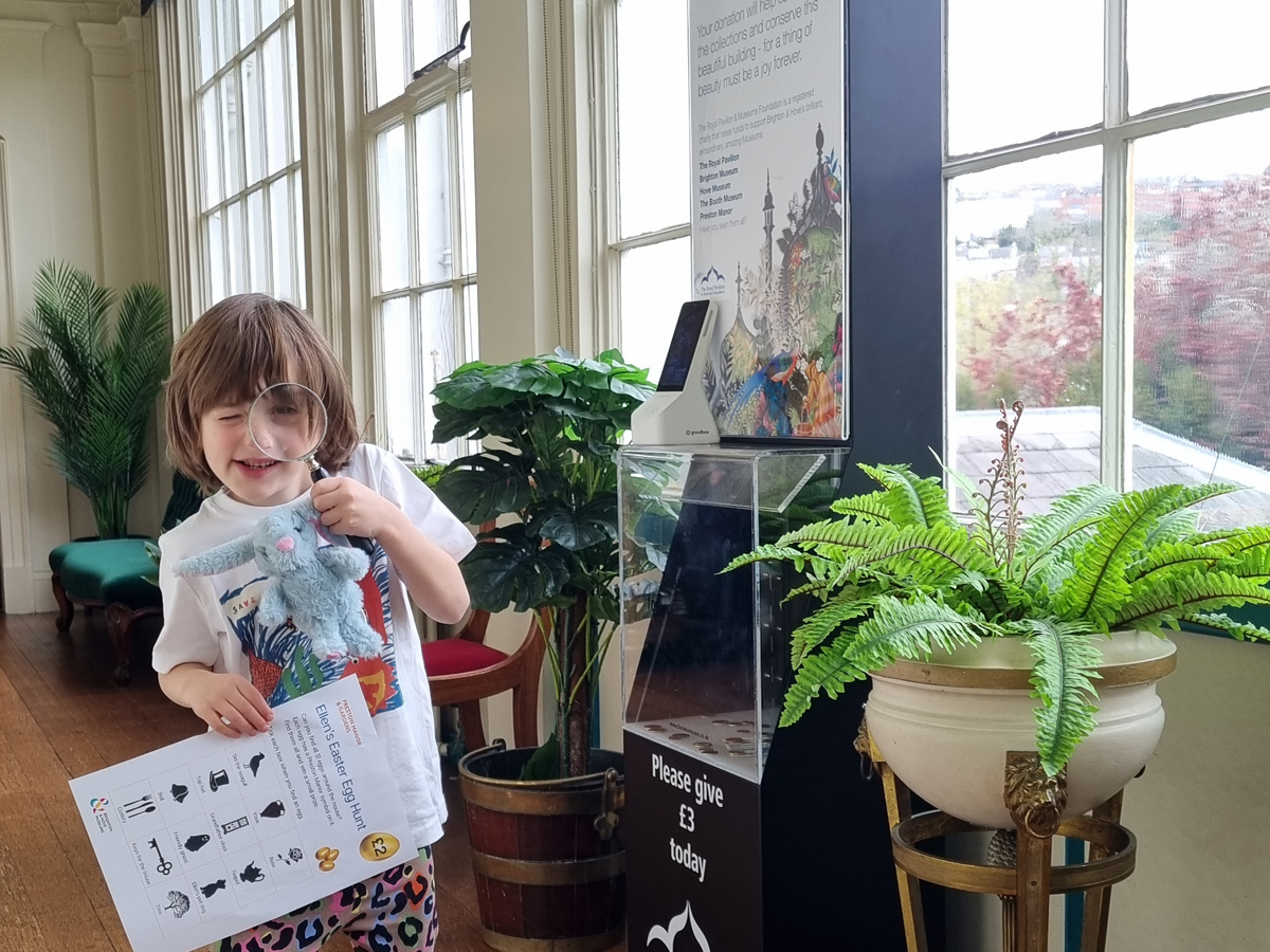 Preston Manor Easter trail 2026. A child stands in the hall of Preston Manor. In one hand they hold a printed Preston Manor children's trail leaflet. The other hand holds a magnifying glass to their face and a toy bunny.