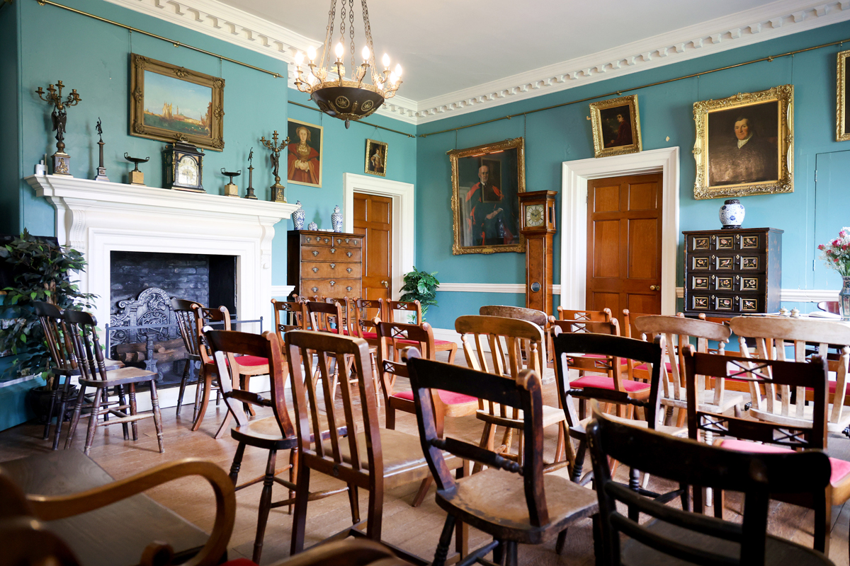 Preston Manor entry hall set for a wedding. The floorspace of the room has been cleared to allow for rows of wooden chairs with red velvet cushions. They face a table with a table runner and a vase of flowers.