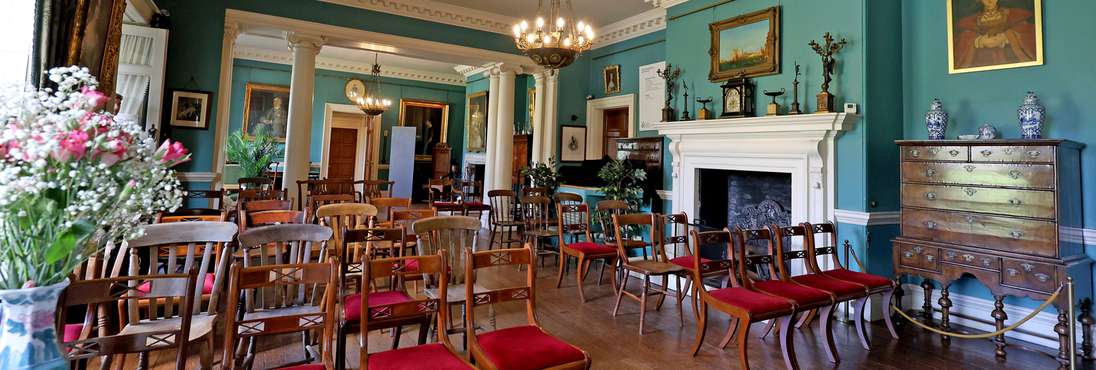 Preston Manor entry hall set for a wedding. The floorspace of the room has been cleared to allow for rows of wooden chairs with red velvet cushions. They face a table with a vase of flowers to the left hand side.