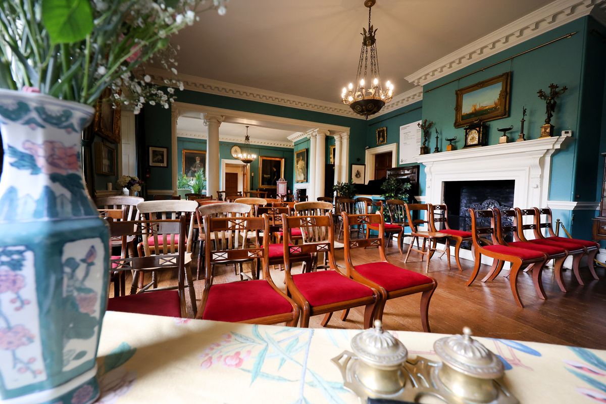 Preston Manor entry hall set for a wedding. The floorspace of the room has been cleared to allow for rows of wooden chairs with red velvet cushions. They face a table with a vase of flowers to the left hand side.