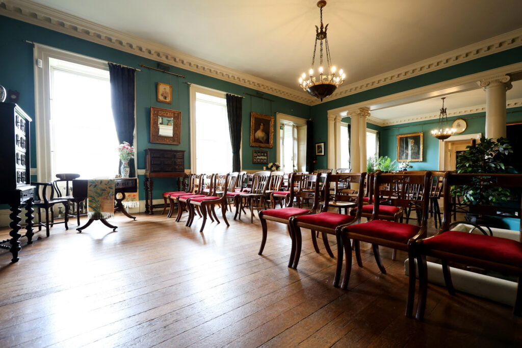 Preston Manor entry hall set for a wedding. The floorspace of the room has been cleared to allow for rows of wooden chairs with red velvet cushions. They face a table with a table runner and a vase of flowers.