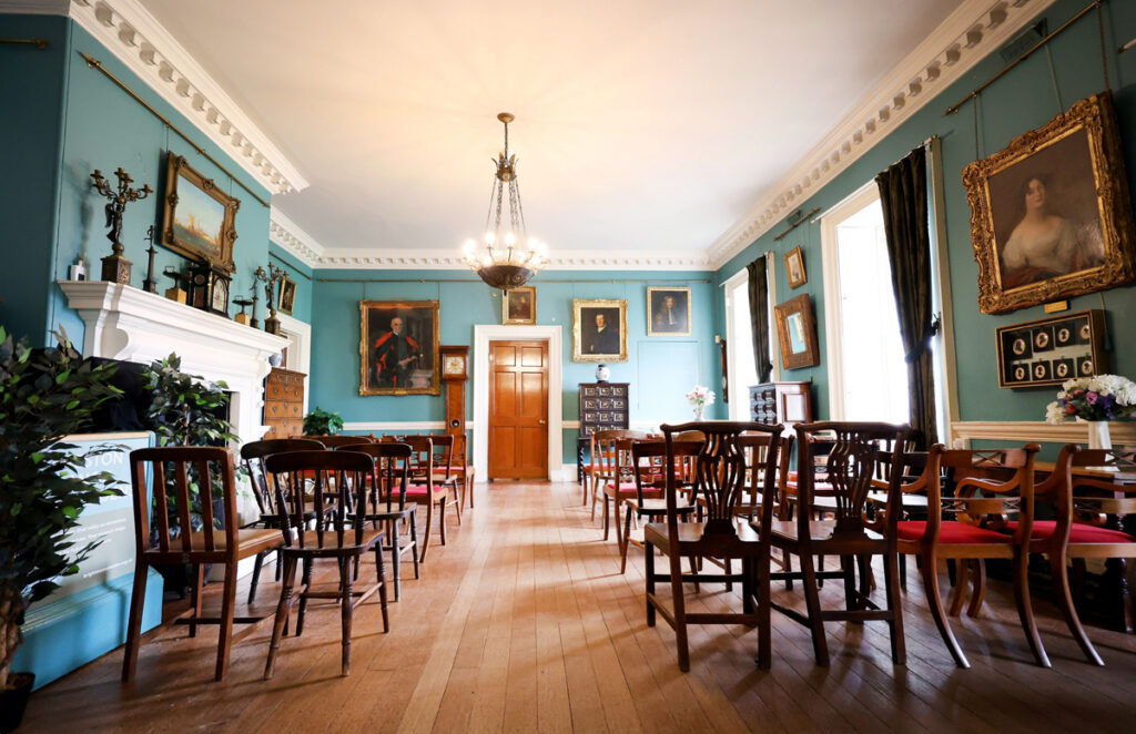 Preston Manor entry hall set for a wedding. The floorspace of the room has been cleared to allow for rows of wooden chairs with red velvet cushions. They face a table with a table runner and a vase of flowers.