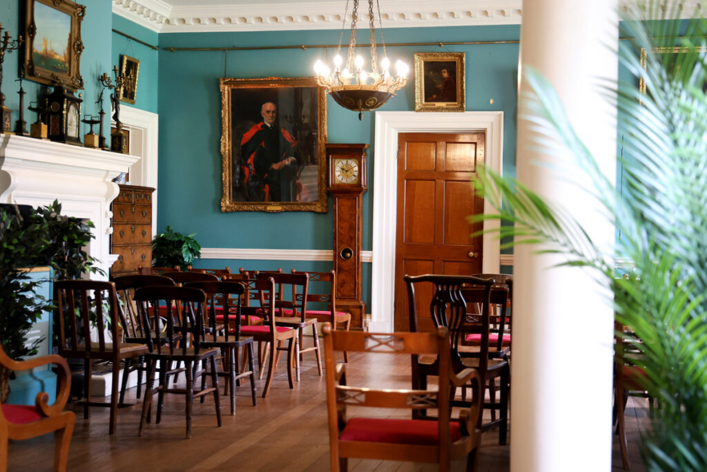 Preston Manor entry hall set for a wedding. The floorspace of the room has been cleared to allow for rows of wooden chairs with red velvet cushions. They face a table with a table runner and a vase of flowers.