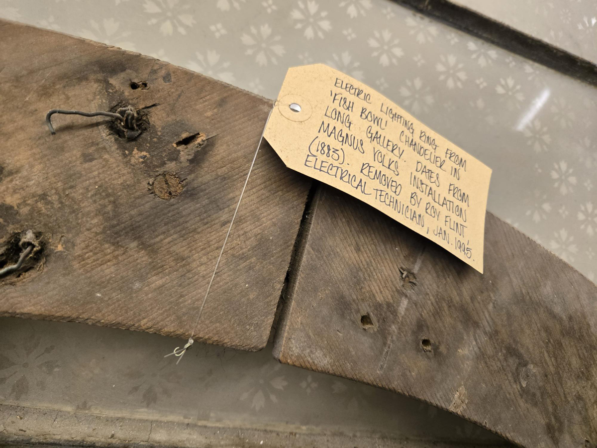 Electrical ring from Fish Bowl Chandelier in Long Gallery. A close up image of an aged wooden ring with a label attached reading the Electrical ring from Fish Bowl Chandelier in Long Gallery, dates from Magnus Volks installations 1883