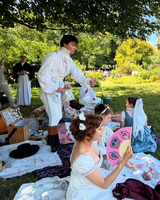 Regency Picnic with Zack Pinsent. A group of people dressed in Regency attire are sitting on blankets in the Royal Pavilion Garden. Some are standing close-by. Picnic hampers and food are laid out on the blanket.