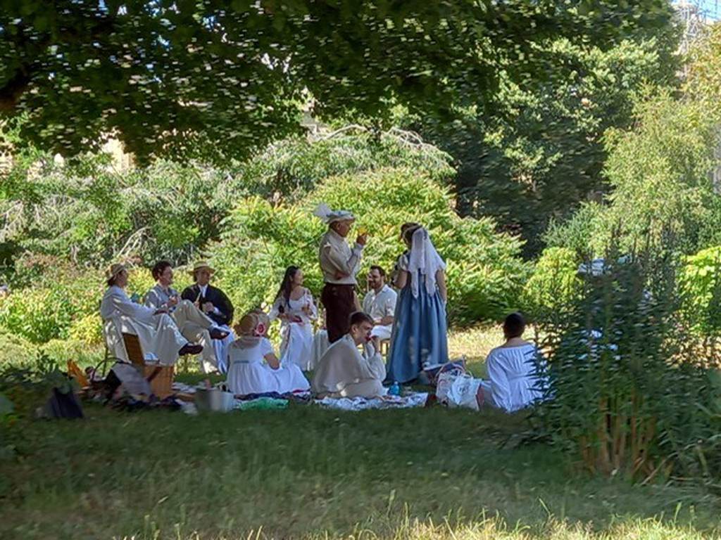 Regency Picnic with Zack Pinsent. A group of people dressed in Regency attire are sitting on blankets in the Royal Pavilion Garden. Picnic hampers and food are laid out on the blanket. The scene is viewed through the trees of the garden.