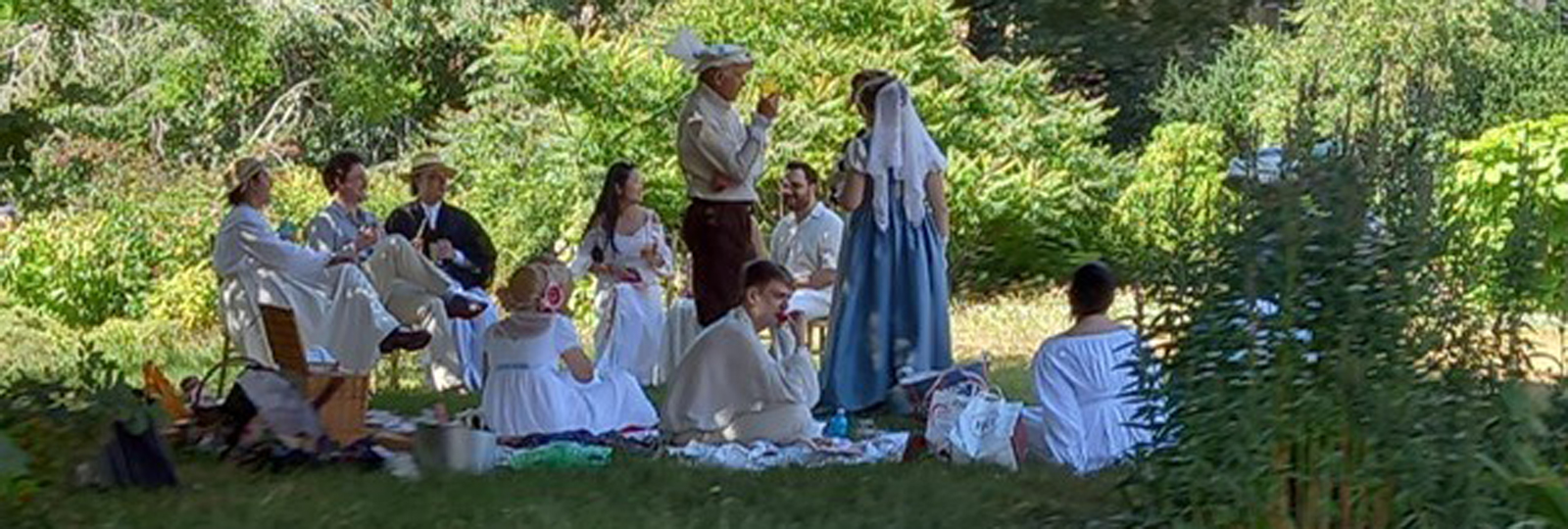 Regency Picnic with Zack Pinsent. A group of people dressed in Regency attire are sitting on blankets in the Royal Pavilion Garden. Picnic hampers and food are laid out on the blanket. The scene is viewed through the trees of the garden.