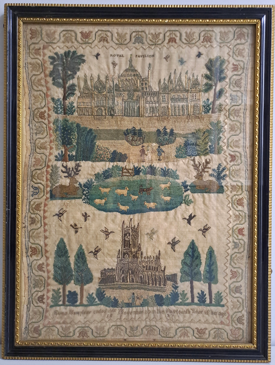 Curator at work, Needlework Samples. A framed piece of needlework. It has a border of flower heads, an image of the Royal Pavilion and Garden at the top. The middle is an animal scene of two seated deer either side of a field with sheep. The lower image is a church with trees either side and a birds flying above.