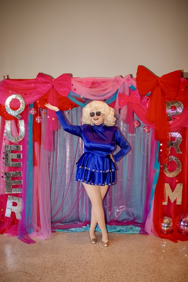 A performer, dressed in a large blonde wig and fabulous short blue dress and sunglasses poses in front of a Queer Prom photo area, adorned with pink bows and ribbons, and netting in various colours.