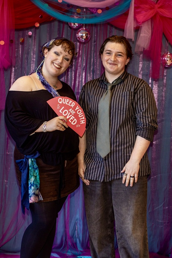 Two guests at the Queer Prom posing for a photo in front of the colourful photograph area, both smiling at the camera