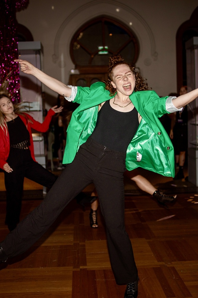 A dancer in black and a flowing green shirt is performing energetically in front of the camera