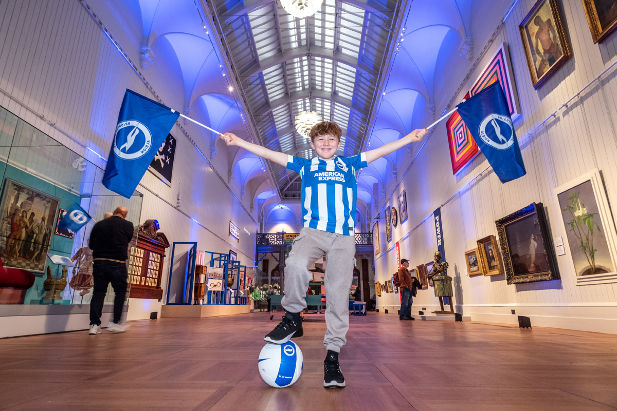 Brighton Museum launch for Brighton and Hove Albion FC's 125th anniversary. A boy stands in the Central Gallery holding a Brighton & Hove Albion flag high in each hand. He wears the football shirt and his right foot rests on a football