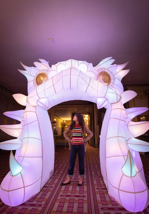 An image of a dragon head inflatable inside a room with a woman standing below. The dragon is illuminated with colourful lights and the woman looks upwards at its head.