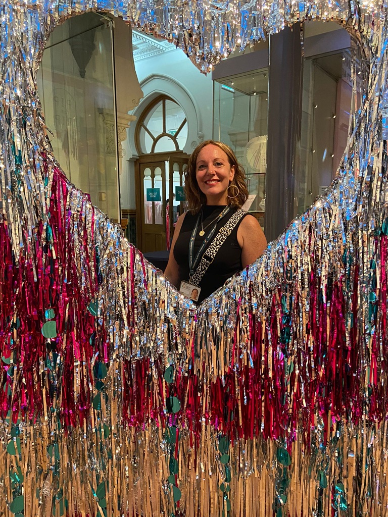 Hazel, a woman with brown hair is smiling at the camera whilst framed in a large tinsel heart.
