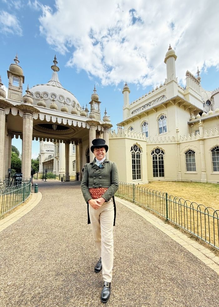 A man dressed in full Regency-style clothing, including a top hat, waistcoat and cream trousers, stands smiling outside the Royal Pavilion in Brighton, with the palace’s distinctive domes and ornate architecture behind him.