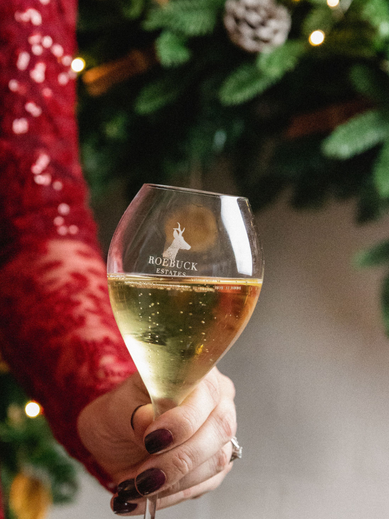 Roebuck Festive image. A woman in a red sparkling top holds a wine glass containing white wine. A tree can be seen in the background.