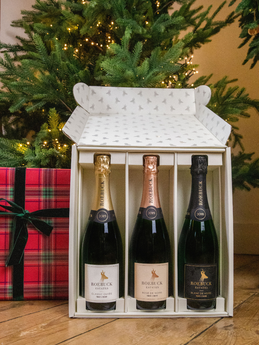 Roebuck Festive image. A box of three bottles of Roebuck wine is placed under a Christmas tree by a wrapped box gift
