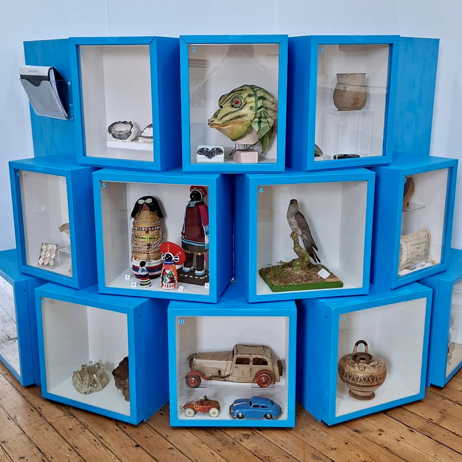What's in the Box? October 2026. Numerous box cases stacked three high and painted bright blue. Each case displays an object from the collections